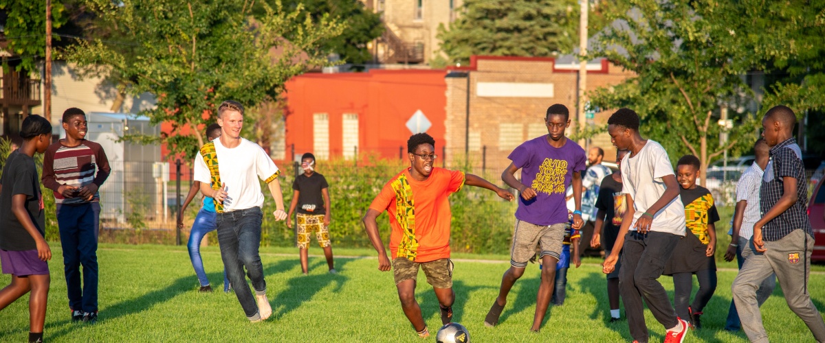 Group of children and teenagers, mostly Black, play soccer at Africa Fest. Their faces are excited, focused, and smiling.